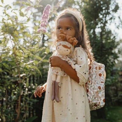 Menina de vestido creme com boneca e mochila floral no jardim.