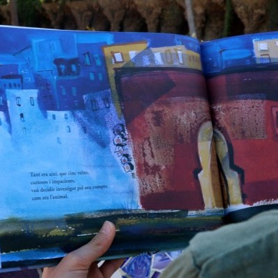 Pessoa a ler livro ilustrado com edifícios coloridos e texto em francês, sentado junto a mosaico colorido.