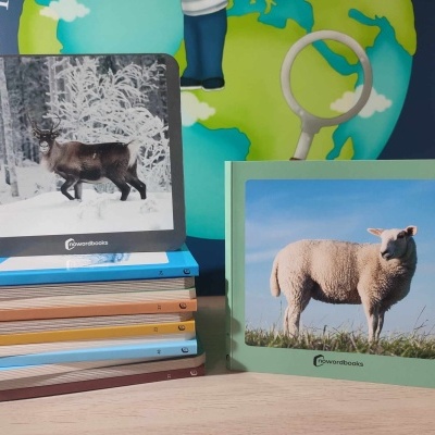 Dois livros infantis com capas rígidas, um com cabra e outro com ovelha, sobre superfície de madeira clara com globo e mapa ao fundo.
