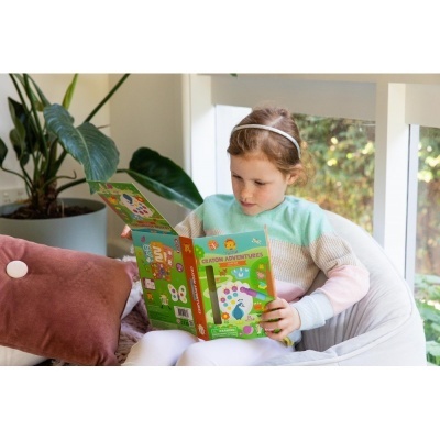 Menina a ler um livro infantil colorido num ambiente interior com planta e almofada