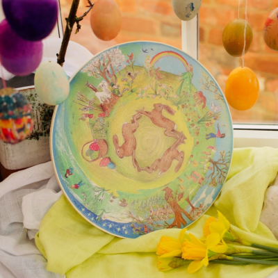 Prato decorativo com ilustração de coelhos e elementos naturais coloridos em fundo de tecido amarelo e ovos de páscoa pendurados