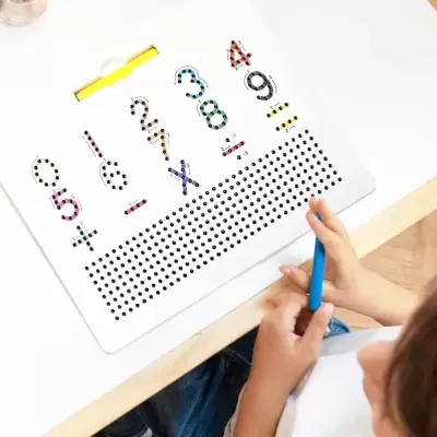 Criança escrevendo em caderno branco com números coloridos e pontos pretos numa mesa.