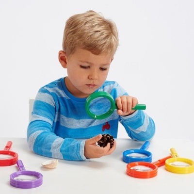 Criança a usar lupa verde para observar objeto, com várias lupas coloridas à sua volta.