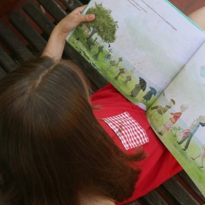 Criança a ler livro ilustrado sentada num banco de madeira, ao ar livre