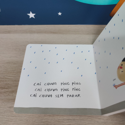 Livro infantil aberto com texto e ilustração de chuva e personagem, sobre mesa de madeira com parede azul e estrelas ao fundo.