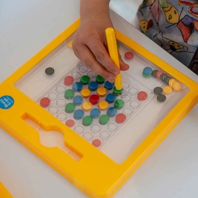 Tabuleiro magnético amarelo com bolinhas coloridas e mão de criança usando vareta amarela