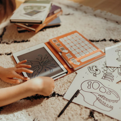 Tablet branco com capa laranja e desenhos a negro, folhas com desenhos de dinossauros ao redor num tapete.