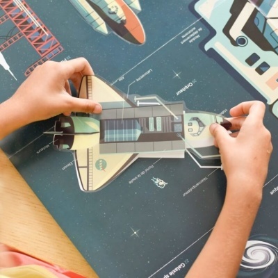 Adulto monta modelo em papel de veículo espacial sobre superfície ilustrada com temas espaciais