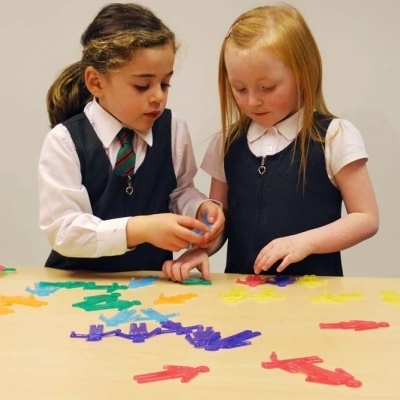Duas meninas com uniformes escolares a brincar com peças coloridas em forma de bonecos numa mesa de madeira.