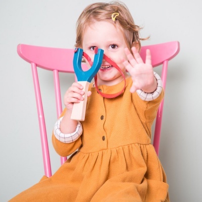 Menina pequena sentada numa cadeira rosa, a vestir vestido amarelo mostarda, segurando funda azul com alça vermelha