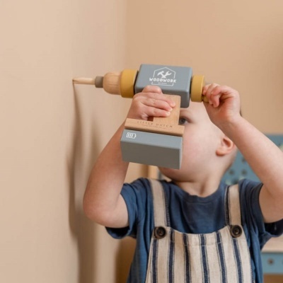Ferramenta de brinquedo em madeira e plástico cinza e amarelo na mão de criança junto a parede bege