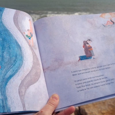 Livro infantil aberto com ilustrações de praia e texto em inglês, segurado com mar e rochas ao fundo.