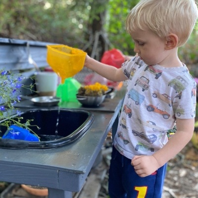 Criança loira a brincar com funil amarelo numa pia preta ao ar livre