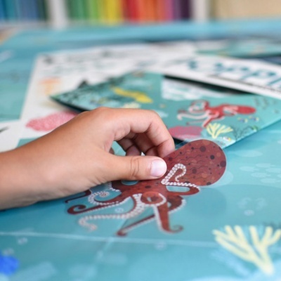 Mão infantil segurando figura de polvo castanho sobre superfície azul com desenhos submarinos.