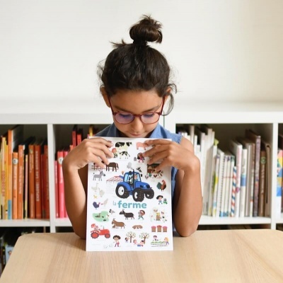 Menina com óculos segura revista colorida com animais e tratores junto a estante de livros