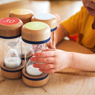 Ampulhetas de madeira e vidro em cores diferentes sobre mesa de madeira com criança ao lado