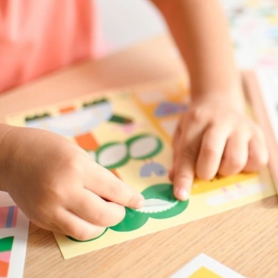 Mãos infantis a aplicar autocolantes verdes numa folha amarela sobre mesa de madeira