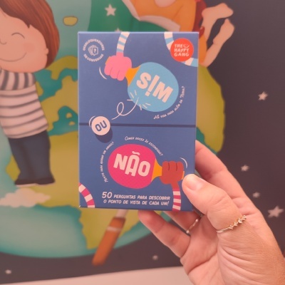 Caixa azul do jogo 'SIM OU NÃO' com ilustrações e texto em português sendo segurada por uma mão feminina com anel, fundo colorido com desenho de crianças e planeta