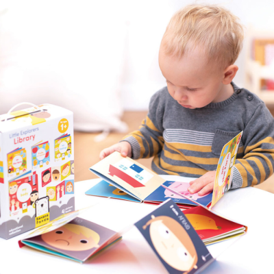 Bebé a ler livros infantis de cartão coloridos sentado ao lado da embalagem do produto Little Explorers Library
