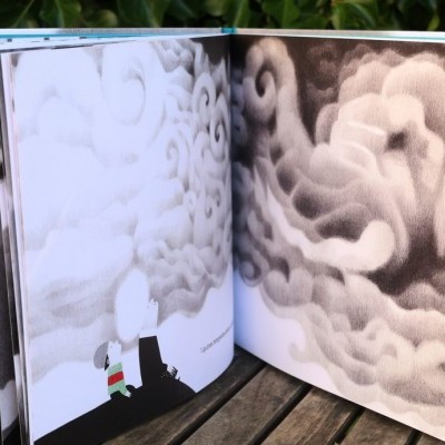 Livro ilustrado aberto com imagens de nuvens e figura humana em mesa de madeira ao ar livre