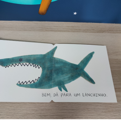 Livro infantil aberto com desenho de tubarão azul e texto em português.