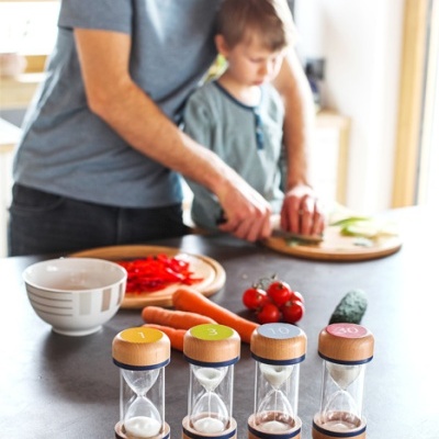 Quatro ampulhetas de madeira e areia em bancada de cozinha com homem e criança a cortar legumes ao fundo