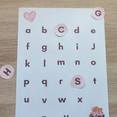 Folha com alfabeto em letras minúsculas e círculos rosas em algumas letras, coração vermelho e bolo no canto inferior direito.