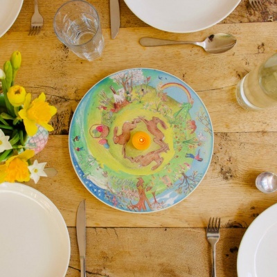 Prato decorativo colorido sobre mesa de madeira com talheres, copos e flores amarelas