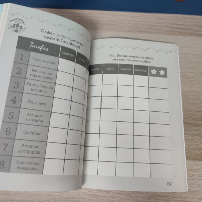 Livro aberto com tabela de tarefas domésticas e dias da semana em português sobre mesa clara.