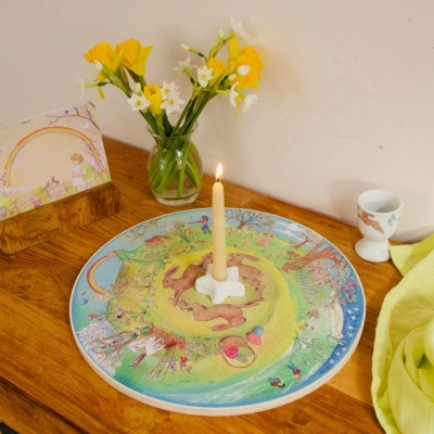 Prato decorativo colorido com castiçal e vela acesa numa mesa de madeira com flores e objetos ao redor