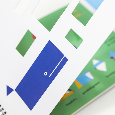 Livro de actividades com páginas recortadas, ilustrando peixe azul e formas geométricas coloridas