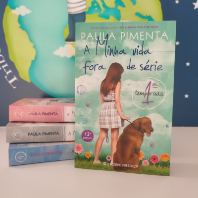 Capa do livro 'A Minha Vida Fora de Série 1ª Temporada' de Paula Pimenta com rapariga e cão, pilha de livros ao lado, fundo com globo terrestre.