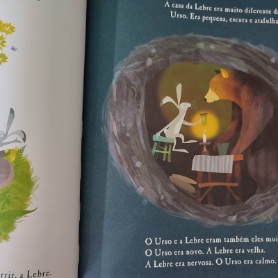 Ilustração de livro infantil com lebre e urso numa toca com vela e texto em português.