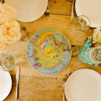 Mesa posta com pratos, copos, talheres, prato decorativo colorido e bule azul decorativo com flores.