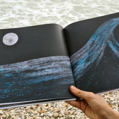 Livro aberto com ilustrações de onda e lua, mão segurando, praia de pedras e mar ao fundo