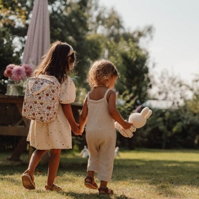 Duas crianças de costas a caminhar num jardim, uma com vestido e mochila estampada, a outra com macacão e boneco de peluche.