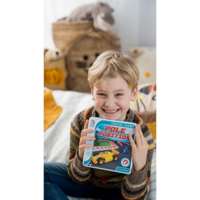 Menino loiro sorridente segurando caixa do jogo de viagem 'Pole Position' sentado na cama