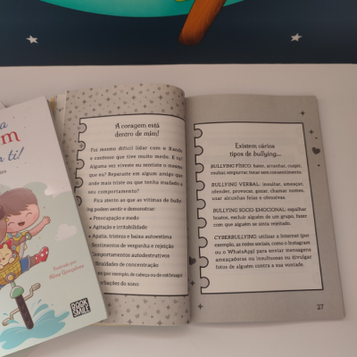 Livro aberto com texto em português sobre bullying em páginas 26 e 27, fundo cinza claro com estrelas