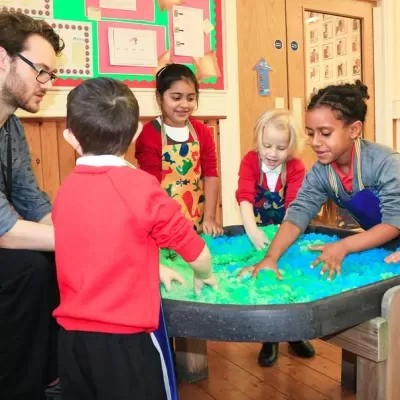 Crianças e adulto em volta de mesa sensorial com material azul e verde, aventais coloridos, sala com madeira e cartazes coloridos.