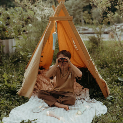 Criança sentada numa tenda amarela a olhar através de binóculos pretos num espaço ao ar livre verde