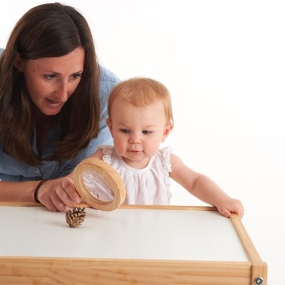 Criança e mulher observam pinha com lente de aumento numa mesa branca com borda de madeira
