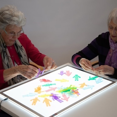 Duas mulheres idosas manuseiam recortes coloridos de figuras humanas sobre mesa iluminada.