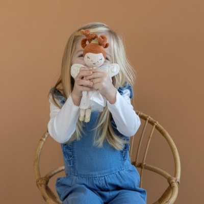 Criança loira segurando boneca de pano com vestido branco sentada em cadeira de vime