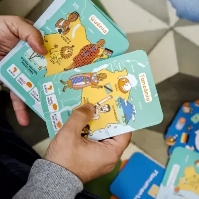 Cartas ilustradas sobre África com texto em português, seguradas por mãos sobre chão de azulejos.