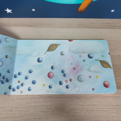 Livro infantil aberto com ilustração colorida de bolas, nuvens, folhas e partículas coloridas sobre fundo azul claro