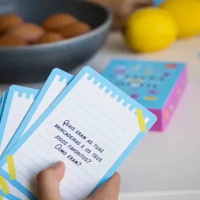 Cartas de jogo azuis com texto em português, mão a segurá-las, tigela com ovos e limões ao fundo