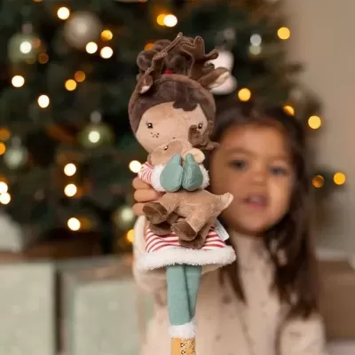 Boneca de peluche com chifres de rena, roupa listrada e botas coloridas, com árvore de Natal ao fundo