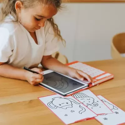 Criança a desenhar num tablet com capa laranja numa mesa de madeira, várias folhas de desenhos para colorir ao lado, fundo com armário branco e decoração natural.