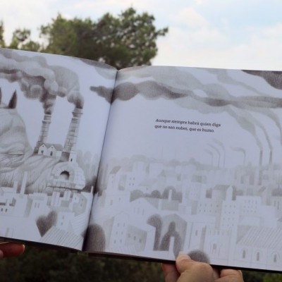 Livro aberto com ilustrações a preto e branco de cidade industrial e mãos segurando-o.