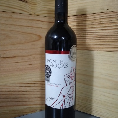 Garrafa de vinho tinto Fonte das Moças com rótulo branco e vermelho numa superfície cinzenta com fundo de madeira.